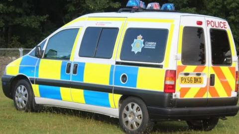 Side view of a police van with blue and yellow markings, parked up in a grassy area. The rear side window has a Cumbria Constabulary logo.