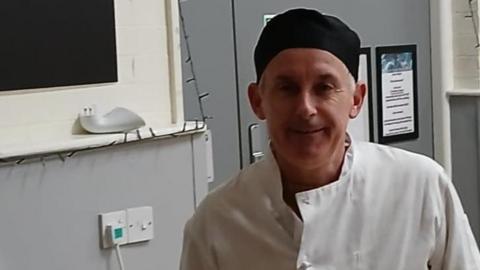 Paul Cleasby smiles at the camera while wearing a chef uniform. He wear a black hat and a white chef's apron. 