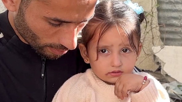 Julia Abu Warda, aged three, sits on her father's knee, as he looks down. There is a wary expression in her brown eyes. She is dressed in a peach-coloured jumper, with her hair in two buns tied with blue bobbles.  