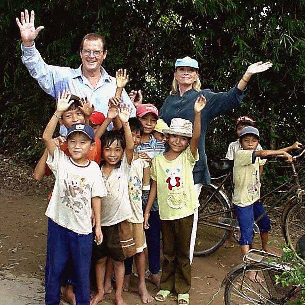 Sir Roger Moore and his fourth wife Kristina are posing for a picture with 10 children in Vietnam. They are all waving at the camera and smiling. Sir Roger is 75 and is wearing glasses and a checked shirt. His wife Kristina is wearing a grey jumper and has a blue baseball cap on her head. One child is sitting astride a bicycle, while the others, all dressed in t-shirts and shorts or trousers, are standing in front of Sir Roger and Kristina looking happy.