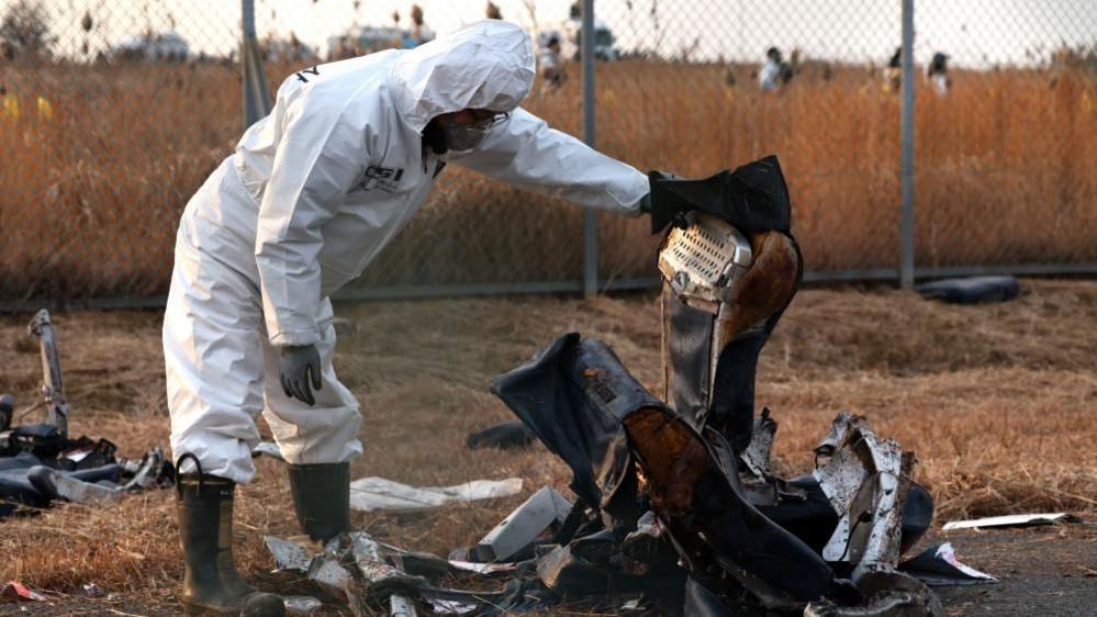 A member of Korea Crime Scene Investigation in a white hazmat suit inspects the wreckage of the Jeju Air aircraft at Muan International Airport. They are holding up what appears to be the remains of a plane seat