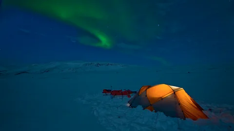 An illuminated tent in a dark snowy landscape with the Northern Lights above (Credit: Getty Images)