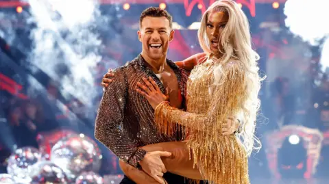 Tayce dancing with Kai Widdrington on Strictly. Tayce is wearing a short gold fitted dress and has long blonde hair. Kai is wearing a sheer black and silver shirt and black trousers and there is a Christmas tree in the background