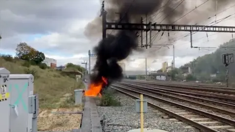 A fire near railway tracks. It is small but has thick black smoke coming from the bright orange flames.