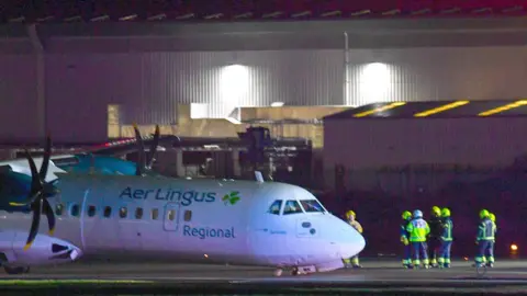 An Aer Lingus plane on the runway at Belfast City Airport.  Emergency crews in high viz clothing are standing close to the plane which appears to have suffered a collapse of its nose wheel.