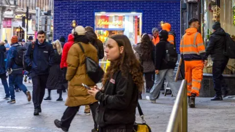 Getty Images A woman stands with a mobile phone in her hands while people are commuting to work on a winter's day