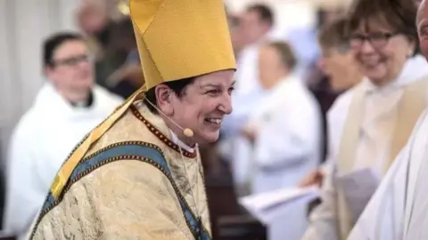 Bishop Anne Dyer, a smiling woman wearing golden-coloured religious clothing surrounded by other members of the clergy.