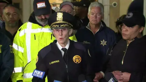 Superintendent Anne Kirkpatrick flanked by several police officers speaking into press microphones