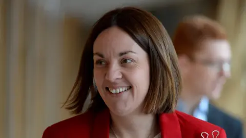 Getty Images A smiling Kezia Dugdale pictured in the lobby of the Scottish parliament building in Edinburgh. She is wearing a white top and a red jacket with a gold brooch. 