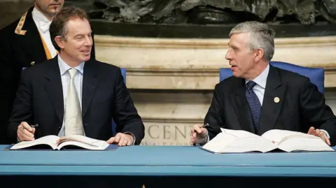 Getty Images Tony Blair and Jack Straw sit at a table with large books in front of them and they are holding pens.