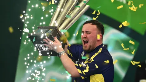 Luke Littler wearing a dark blue and yellow top lifting a gold PDC trophy as yellow confetti goes off around him