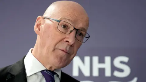 A bald man with glasses, wearing a dark suit and purple tie, in front of a purple background with an NHS Scotland logo in a close-up shot. 