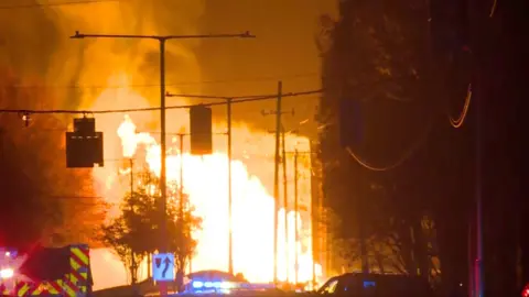 Flames as tall at street lamps on a dark street 