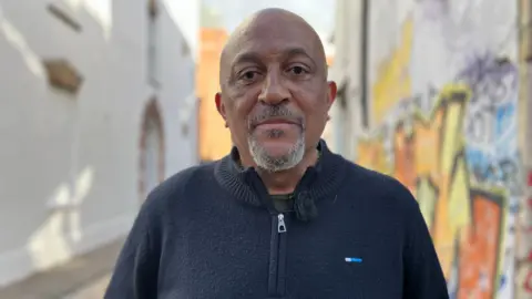 Desmond Brown. He is bald and has a grey goatee. He is wearing a navy blue fleece and is standing in a street, near a wall covered in multicolour graffiti. Desmond is looking at the camera and smiling. 