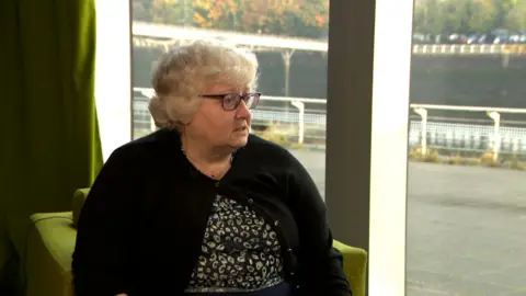 Laura Schosloff wears a flowery top and black cardigan while sitting in an office with the River Clyde in the background