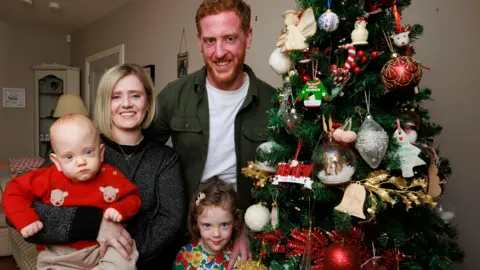 PA Media Lindsay Ace, Matthew Ace, four-year-old Aine Ace and Iarla Ace stand beside a christmas tree. Lindsay is smiling and holding Iarla. Matthew Ace has his hand on the shoulder of Aine as they smile.