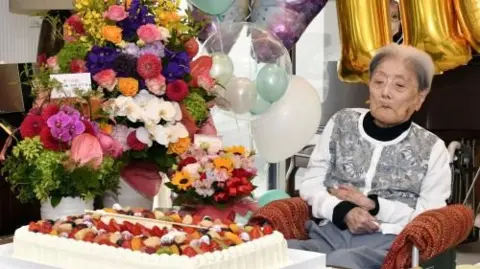 Tomiko Itooka celebrates her 116th birthday. She sits in front of a large white cake topped with fruit and a bouquet of flowers.