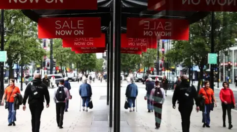 Lots of people walking in front of the mirrored front of a shop with red banners flying over their heads saying "sale now on"