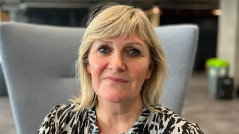 Angela MacVicar, with shoulder length blonde hair and wearing a patterned black and white blouse, sitting on a grey chair as she looks straight at the camera