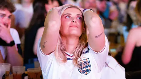 A female England football fan looks at a big screen at the Propyard venue in Bristol, with her hands behind her head. She has blonde hair and is wearing a white England replica shirt.