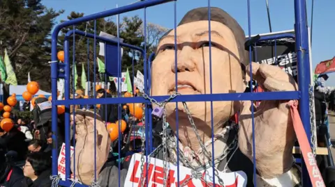 Getty Images A sculpture of South Korean president Yoon Suk Yeol is draped in chains inside a blue cage and surrounded by protesters in the street