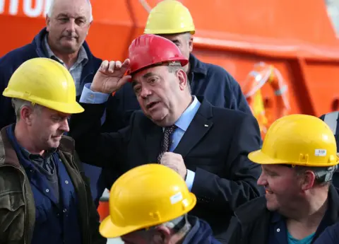 PA Media Alex Salmond wearing a red helmet, surrounded by yellow helmetted workers