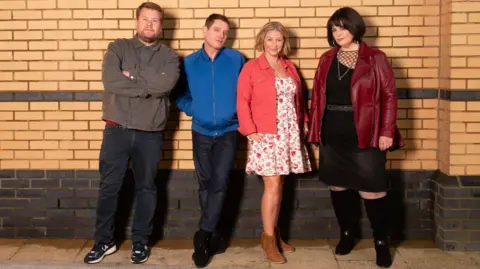 Four members of the cast of Gavin and Stacey reunite for an official cast photo: Smithy, Gavin, Stacey and Nessa. They are stood against a brick wall. Smithy (played by James Corden) is wearing a grey jacket and blue jeans and has his arms crossed, Gavin (played by Mathew Horne) is next to him, wearing a blue jacket with his hands in the pockets and blue jeans. Next to him is Stacey (played by Joanna Page), she is wearing brown cowboy boots, a white floral dress and a pink jacket, and she is stood next to Nessa (played by Ruth Jones), who is wearing a burgundy leather jacket and a black dress with long black boots.