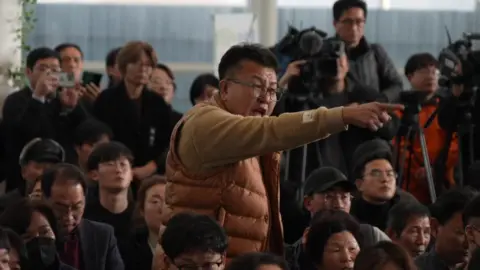 A man, standing in an airport waiting room, speaking and pointing his finger at someone off camera