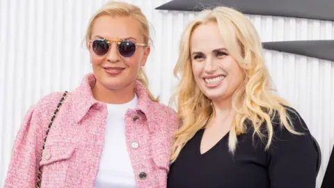 Ramona Agruma and Rebel Wilson attend the Women's Single Finals of the US Open. Romana is wearing sunglasses and has her blonde hair tied back. She is wearing a pink jacket with a white t-shirt underneath. Rebel is smiling and has her blonde curly hair down. She is wearing a black dress.