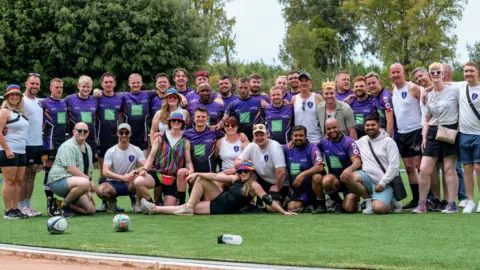 A group shot of about 35 members of the Colchester Kings. There are two lines of players, most in purple and black kits, one standing, the other kneeling. The players and their supporters are all smiling. The picture has been taken on a rugby pitch. 