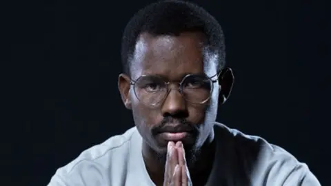 Nelson Amenya, in a white top, golds his hands in front of him in a prayer position as he stares into the camera.