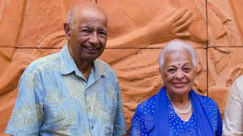 Gulab Watumull seen wearing a blue, patterned Aloha shirt with his wife Indru who is wearing a blue kurta with a pattern of white dots and a pearl necklace, in 2012 