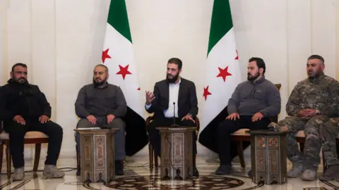 Ahmed al-Sharaa wearing a dark suit and white shirt sitting in front of two Syrian flags with with three men in dark clothing and one in military uniform from Syria's new joint forces.