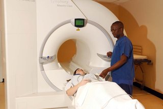 Person lying on a flat bed being moved into an MRI scanner by a radiographer operating the controls