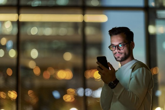 Man having video chat in office building at night with lights twinkling outside the windows behind him.