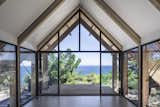 A double-glazed end wall with two doors frames stunning views of the Aegean Sea. A deep roof overhang shelters the outdoor terrace.