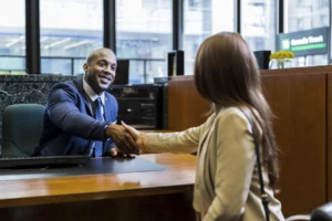 Two people sitting at a desk across from each other in front of a window shaking hands after an interview.