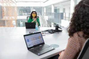 Two female employees wearing face masks and talking in a conference room while social distancing. Hybrid Workplace collection. Keywords: Hybrid Workplace, Hybrid Worksite, Return to Workplace, Return to Work, Return to Worksite, Return to Office, Return to Campus, Reopening, Back to work, COVID-19, COVID-19 policies, COVID, Coronavirus, Pandemic, Health, Health and Safety, Face Covering, Face Mask, Masks, Masks at Work, Masks in Workplace, Open Workspace, Open Workspace Policies, Social Distancing, Social Distancing Policies, Social Distance, Physical Distancing.