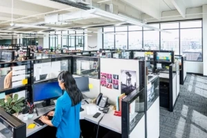 Female call center employee standing at cubicle desk, using desktop computer. Two monitors shown (one with Windows 7 log in screen visible) and a phone on desk. There are several other cubicles in the call center office.