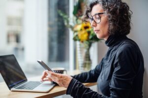 Mature businesswoman sitting at cafe looking at her cell phone while working on laptop computer.