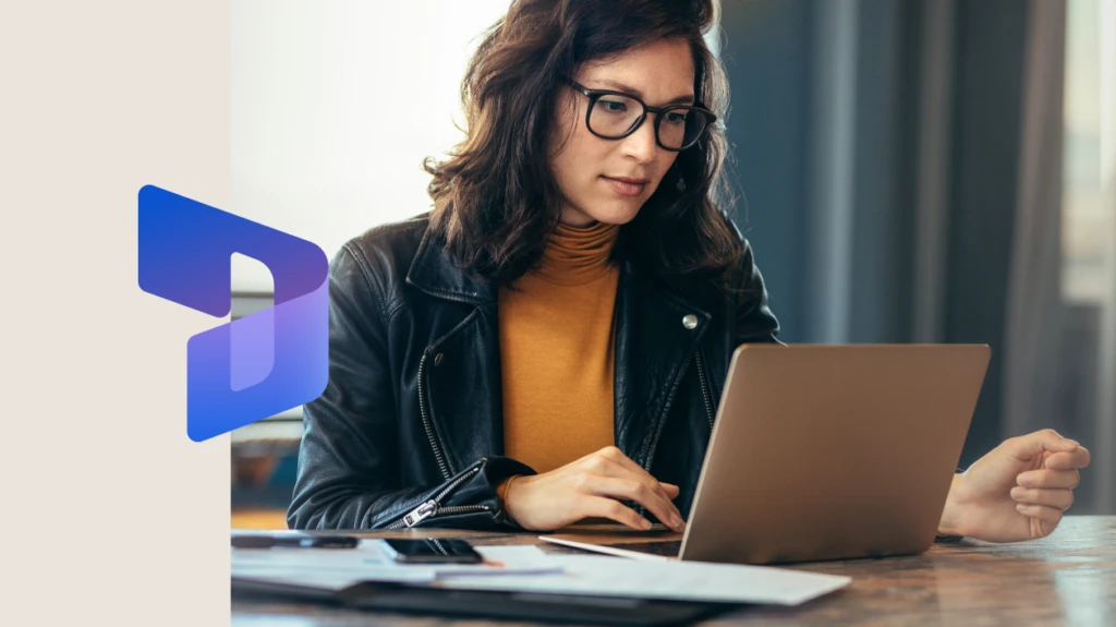 Businesswoman sitting at a table in an office, typing on her laptop with focus. The image is overlaid with the Dynamics 365 icon on the left.