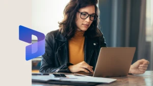 Businesswoman sitting at a table in an office, typing on her laptop with focus. The image is overlaid with the Dynamics 365 icon on the left.