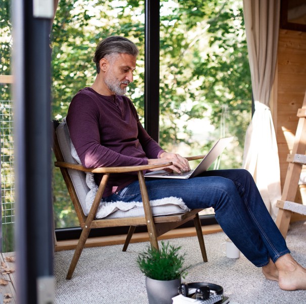 A mature man sits inside a treehouse, using a laptop.