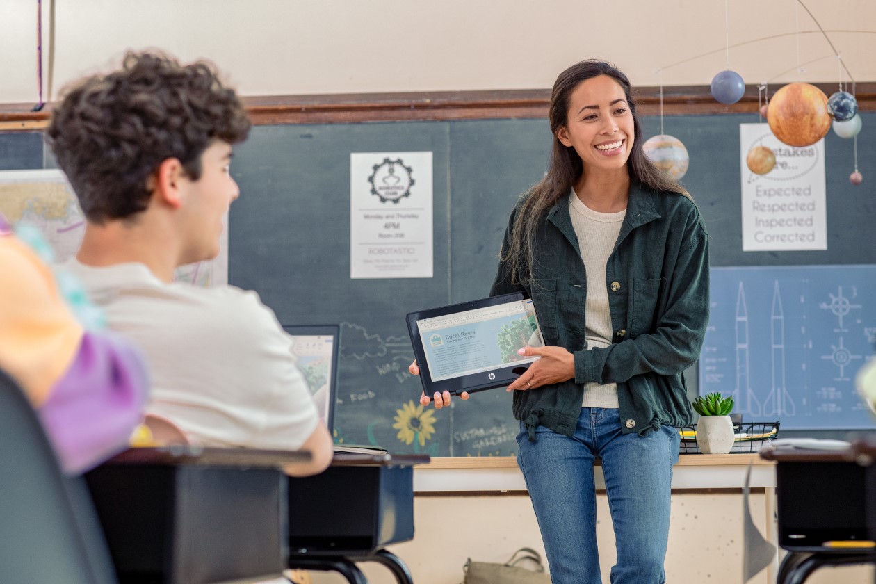 Decorative. A teacher holding a folded laptop in front of students in a classroom.