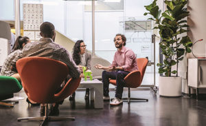 A team meets in a conference room. All workers are turned toward one employee who is giving an explanation.