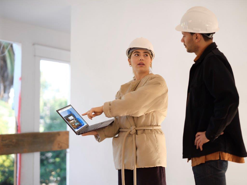 Two frontline workers wearing hard hats reviewing plans on a laptop.