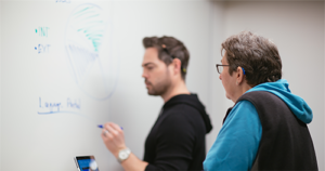 A woman who wears a hearing aid, watches a colleague write on a whiteboard in a conference room.