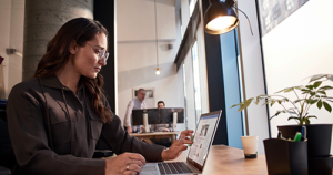 An image of a female tech worker sitting at a table working on her laptop.