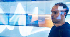 An image of a man in hooded sweater/sweatshirt inside a secure room who is looking at data and a geographic area displayed on a large monitor which is behind glass walls with reflections.