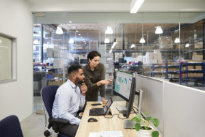 Two adults in an office adjacent to a warehouse setting collaborating while overlooking dual monitors.